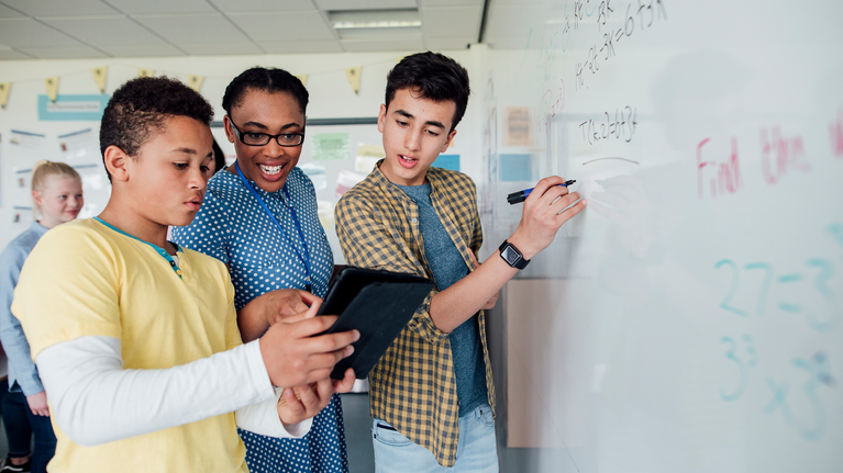 Teenage student writes on a whiteboard whilst another student and teacher are using a tablet device