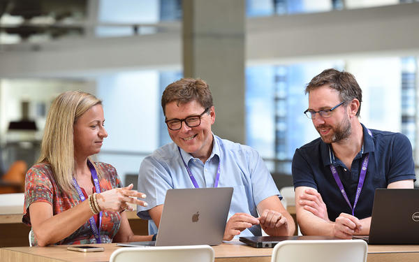 Three Cambridge colleagues sit in front of their laptops smiling while the female colleague points to her screen for her other two male colleagues to look at