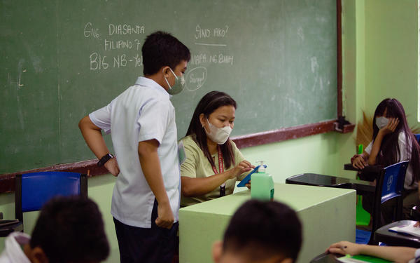 Child approaches teacher at desk in classroom. Both wear protective masks.