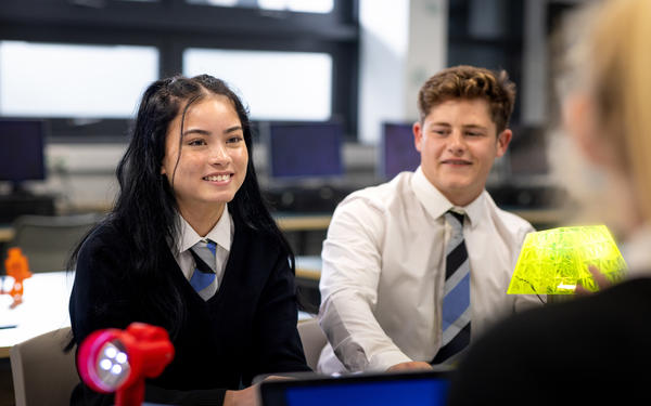 An over-the-shoulder view of two teenage students in their school uniforms having a conversation in the classroom during a lesson in a class in school in the North East of England.