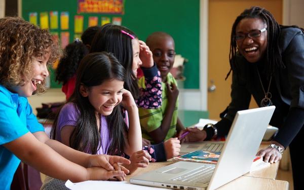 A teacher leaning over and talking to a group of young students who are working at laptops