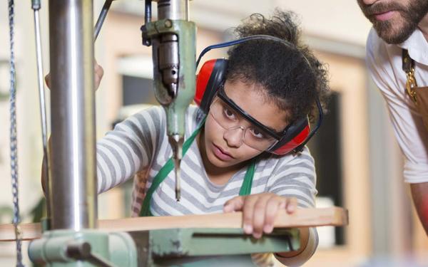 Woodwork student being observed by a teacher while working at a machine, wearing protective goggles and ear defenders