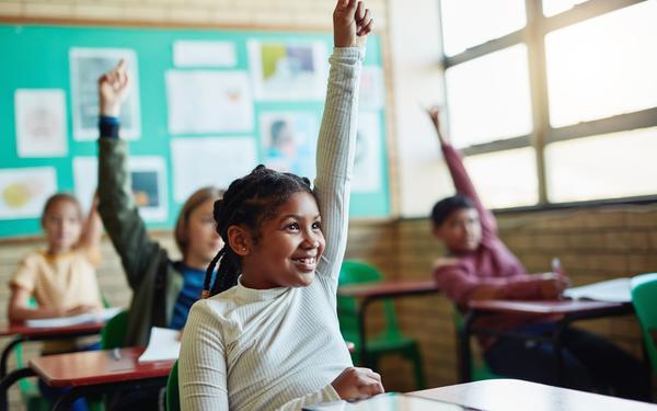 a young student holds her hands up in class while sat behind a desk