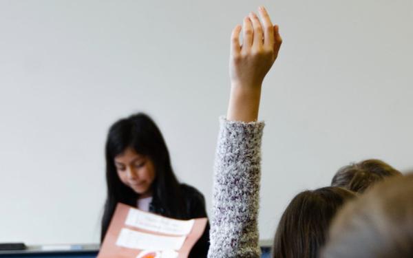 someone holds their hand up in foreground as a young girl stands at front of a class in the background