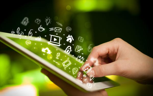 Hand holding a tablet with graphics of app icons floating out of the screen. The other hand is touching the screen of the tablet. Blurred green background.