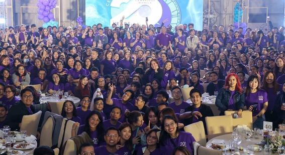 Cambridge's 900 manila colleagues gather together on and off stage in front of a screen for a group photo dressed in Cambridge's purple 20th anniversary t-shirts.