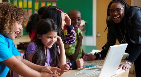 A teacher leaning over and talking to a group of young students who are working at laptops