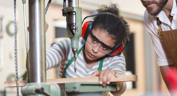 Woodwork student being observed by a teacher while working at a machine, wearing protective goggles and ear defenders