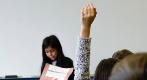 someone holds their hand up in foreground as a young girl stands at front of a class in the background
